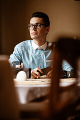 LGBTQ community lifestyle concept. Young homosexual man sits at the table in old-fashioned city cafe. Handsome smiling gay male businessman poses while having a break at lunch