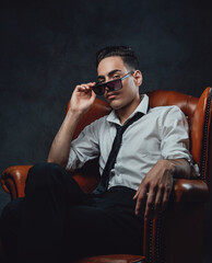 Obraz na płótnie Canvas Young confident man in sunglasses wearing white shirt and black tie sitting on a luxury chair and looking on the camera against black background