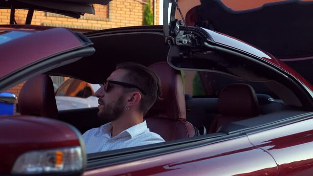 The convertible has a roof. The man is sitting in a convertible. Handsome guy with glasses sitting in the car.