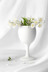 White still life with jasmine flowers in a white wine glass with a background of flowing white fabric.