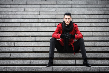 LGBTQ community lifestyle concept. Young homosexual man sits on a stairs. Handsome fashionable gay male model poses in cityscape outdoors. Wears red coat, gloves, and black scarf