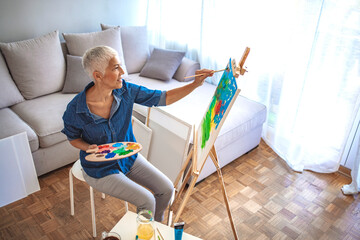 Senior artist working in concentration. Side view portrait of white haired senior woman holding palette painting pictures at easel in art studio standing against windows in sunlight