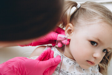 Charming little girl having an ear piercing process with special equipment in a beauty center by a...