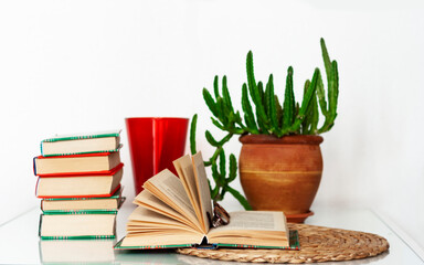 Cozy home interior decor: cup of coffee, stack of books, plants in pots, open book on a wicker stand, pillows on a white table. Distance home education.Quarantine concept of stay home