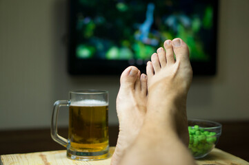 Feet on the table in front of the TV, On the table a mug of beer, a snack in the background of the TV. Rest after work, Friday evening, before weekends, holidays at home alone.