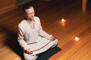 A portrait of an attractive Caucasian man sits in cotton robes in a lotus pose in a dark wooden...