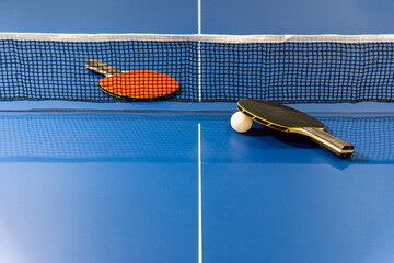 Black and red table tennis racket and a white ball on the blue ping pong table with a net, Two table tennis paddle is a sports competition equipment indoor activity and exercise for background concept - obrazy, fototapety, plakaty
