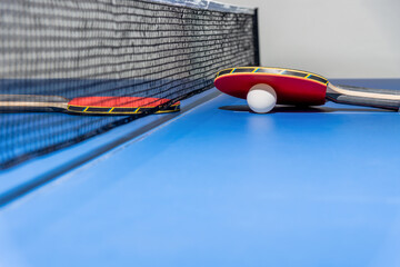 Red table tennis racket and a white ball on the blue ping pong table with a black net, Two table tennis paddle is a sports competition equipment indoor activity and exercise for concept background
