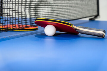 Red table tennis racket and a white ball on the blue ping pong table with a black net, Two table tennis paddle is a sports competition equipment indoor activity and exercise for concept background