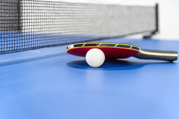 Closeup red table tennis racket and a white ball on the blue ping pong table with a black net,...