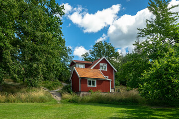 Fototapeta na wymiar Typical village red color Swedish house surrounded by garden trees at summer. Green mowed lawn in front of the building. Beautiful rustic countryside wooden house with tiled roof against blue sky.