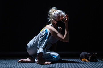 Blonde movement artist in white clothing painted with blue, yellow and black, wearing a black mask, is dancing alone in a darkened studio.