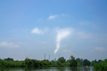 A tall chimney on the horizon near a river smokes into the sky.