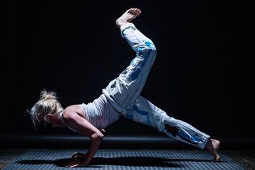 Blonde movement artist in white clothing painted with blue, yellow and black, wearing a black mask, is dancing alone in a darkened studio.