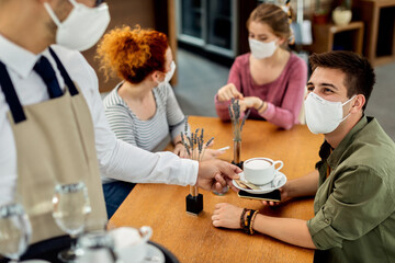 Happy man wearing protective face mask while drinking coffee with friends in a cafe.