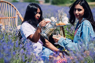 Fototapeta premium Two beautiful indian girls sitting in purple lavender field with decoration and pouring drink on glasses.