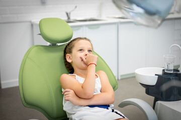 Little girl looks worried with fear closes mouth with handssitting in a dental chair. Visiting dentist with children
