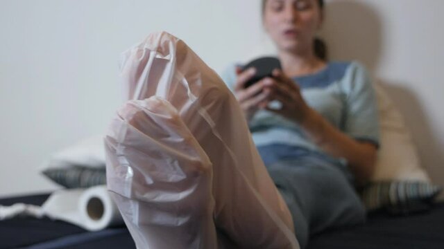 Moisturizing foot mask for dry heels. A woman in a mask on her legs lies on the diva and uses the phone. Self-care during the period of the virus at home