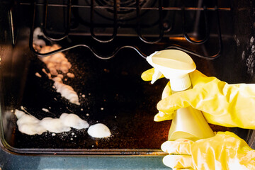 cleaning oven in home kitchen - hands in gloves spray a reagent on dirty bottom of oven