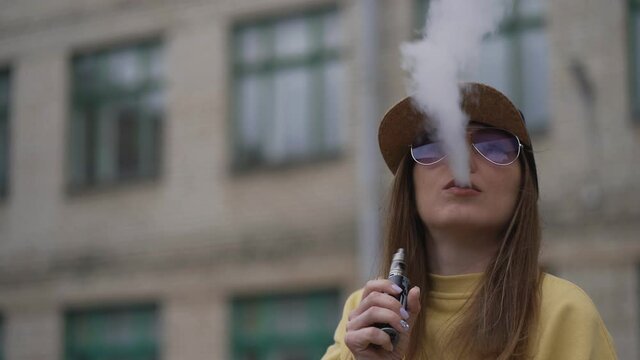 Pretty young hipster woman in black cap yellow hoodie vape ecig. Girl vape popular ecig gadget, vaping device.Happy brunette vaper girl with e-cig. Portrait of smoker female model with electronic