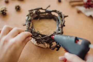 Making rustic christmas candle holder. Hands holding glue gun and making rustic festive decoration from branches, pine cones and cotton on wooden table. Holiday workshop