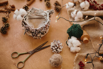 Making rustic christmas candle decor. Christmas candle  holder made of branches, cotton, anise and pine cones on wooden table with festive lights. Holiday workshop.