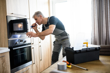 Here for Repairs. Aged repairman in uniform working, examining broken microwave in the kitchen using flashlight. Repair service concept