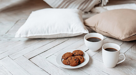 Breakfast in the morning. Two white cups of coffee and chocolate chip cookies stand on a light wooden parquet and pillows lie nearby. Homeliness, day off. Pastel colors.