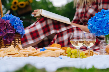 Girl in red checkered dress and hat sitting on white knit picnic blanket reading book and drinking wine. Summer picnic on sunny day with bread, fruit, bouquet hydrangea flowers