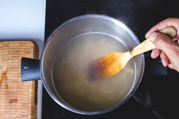 Mixing and cooking with a wooden spoon boiling water in an inox pot on a domestic kitchen