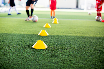selective focus to yellow cone marker is soccer training equipment on green artificial turf with blurry kid players training background. Material for trainning class of football academy.