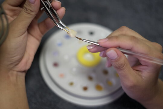 Dental technician applies ceramic material to the crown using tweezers and brush on dental palette background .Dental technology close-up