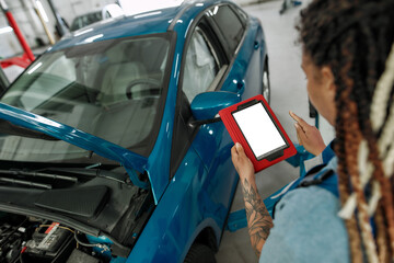Quality Trusted. Young african american woman, professional female mechanic looking at the blank screen of tablet pc, standing near car with open hood at auto repair shop