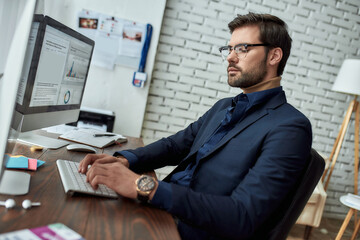 Fototapeta na wymiar Side view of a financial analyst wearing eyeglasses working with statistical data while sitting at his workplace in the modern office