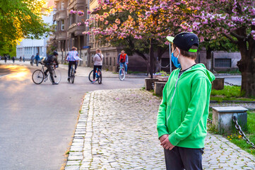 A lonely boy in a protective mask watches a group of teenagers on bicycles. Quarantine coronavirus covid-19. Outcast in the company. Sad lonely teen looks at teenagers with envy