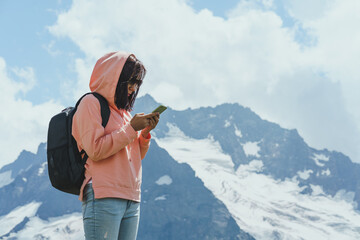 Female traveler stands with smartphone in mountains. Woman tourist browsing mobile phone against cloudy sky on sunny day in mountainous terrain.