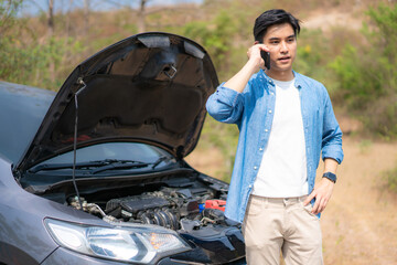 Asian young unhappy man talking on a cell phone  in front of the open hood  broken down car On Country Road Phoning For Help. Broken Car On The Road in During journey..