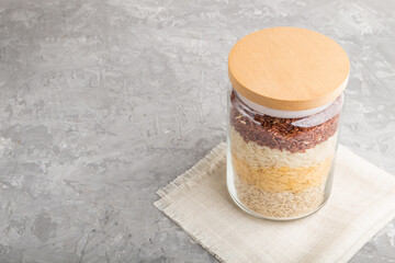 glass jar with different kinds of rice poured in layers on a gray concrete background. side view, copy space.