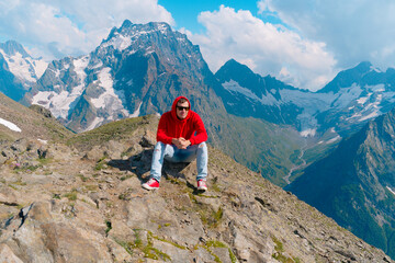 Young man in sunglasses sitting on mountain in sunny weather. Adult male in red hoodie with hood enjoying beautiful view in mountainous area.