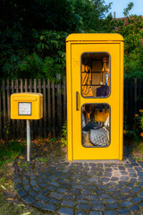 Ein gelber Briefkasten steht neben einer gelben Telefonzelle,  in der, weil sie nicht mehr funktionstauglich ist, Reitutensilien ausgestellt sind, im Hintergrund ein Holzzaun vor Bäumen und Büschen