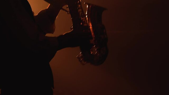 Silhouette a young stylish guy plays the golden shiny saxophone in the yellow spotlights on stage. Dark studio with smoke and neon lighting. Accent on the saxophone. Close up.