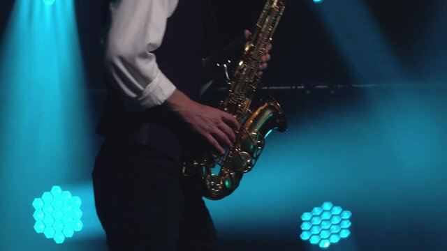 A young retro stylish guy plays on the golden shiny saxophone in the turquoise spotlights on stage. Dark studio with smoke and neon lighting. Hands and saxophone close up. Side view.