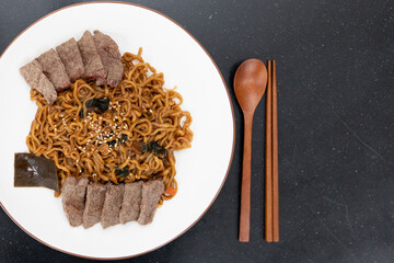 Top view image of korean black bean noodles served with sliced meat with wooden eating utensils.