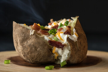 Large steaming baked potato topped with bits of bacon, chopped green onion, cheese and sour cream ready for eating.