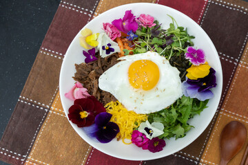 Colorful edible flowers with fried egg and meat salad on a bowl with wooden eating utensils