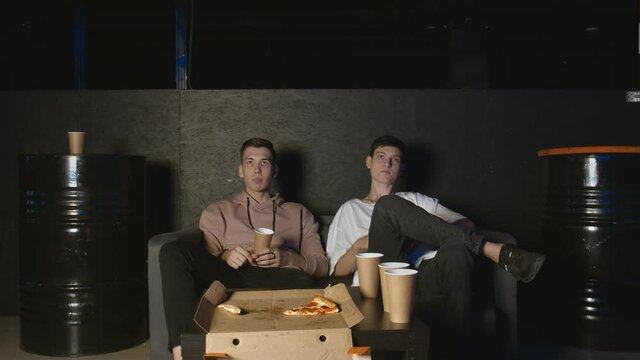 Two best friends watching television together on couch, communicating, eating popcorn and pizza