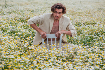 Tall handsome man sitting on a white chair in camomile flowers field