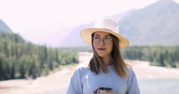Young woman in hat is drinking a tea in mountains at summer sunny day, front view. She is looking at camera and smiling and admiring scenery view in nature park. Traveling, vacation, journey concept.