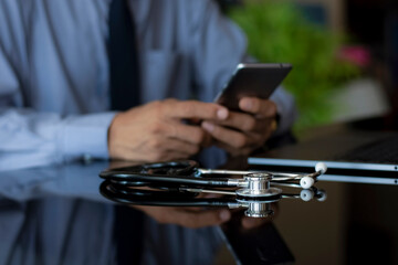 Smart male doctor hand holding and using mobile smart phone with stethoscope and laptop computer on the desk in medical room at clinic or hospital. Medic tech, online medical concept.