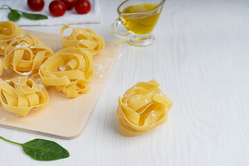 Closeup side view of raw organic italian fettucine pasta powdered by flour on cutting board surrounded by spinach, tomatoes and olive oil gravy boat on white wooden background. Image with copy space